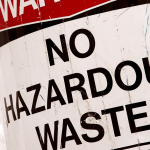Weathered outdoor sign reading 'NO HAZARDOUS WASTE' with a red warning strip at the top.