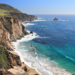 Cliffside coastline with rugged yellow and green cliffs meeting turquoise ocean, a sandy beach tucked below, and a clear blue sky.