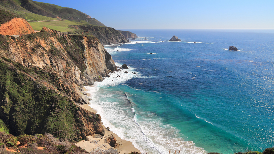Cliffside coastline with rugged yellow and green cliffs meeting turquoise ocean, a sandy beach tucked below, and a clear blue sky.