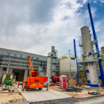 Industrial construction site with large processing towers and blue pipes against a cloudy sky, foreground showing an orange crane and scattered building materials.